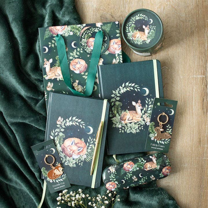 A collection of items featuring woodland critters, including an enamel keychain of a baby deer named Felicity Fawn. Also pictured for aesthetics is a fox keychain, fox and fawn notebooks, eyeglass holders, candle, and tote bag.