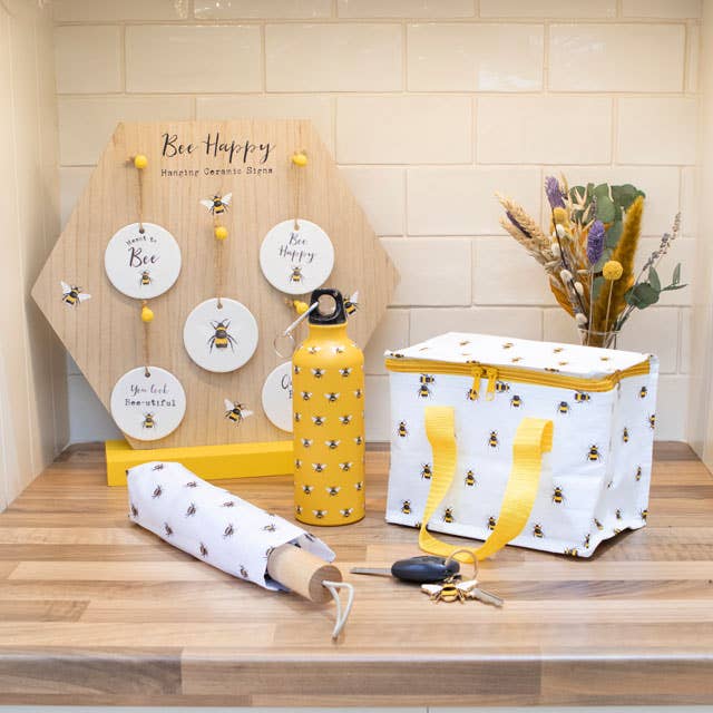 A collection of bee print items, including an insulated lunch tote with yellow handles and zipper closure. Photo also shows umbrella, keychain, ornaments, and water bottle with bees.