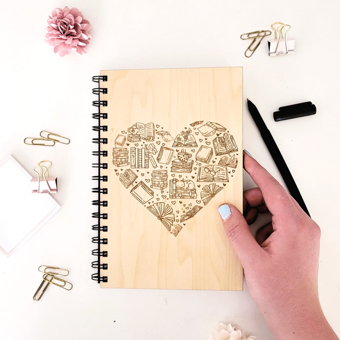A wood journal with a laser-engraved design in the shape of a heart featuring bookish icons.